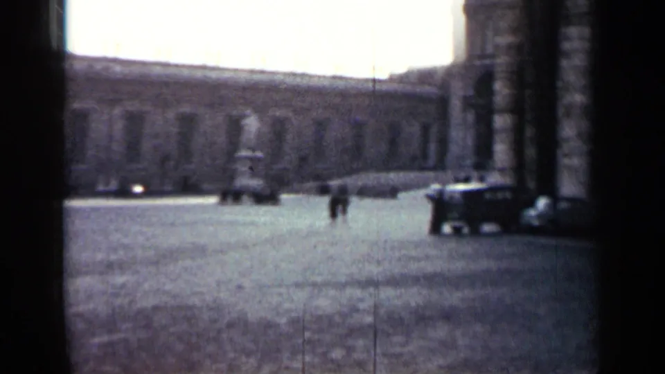 1962 Cuba street frame with distant pedestrians crossing an open cobblestone area beside cars and a large historic facade.