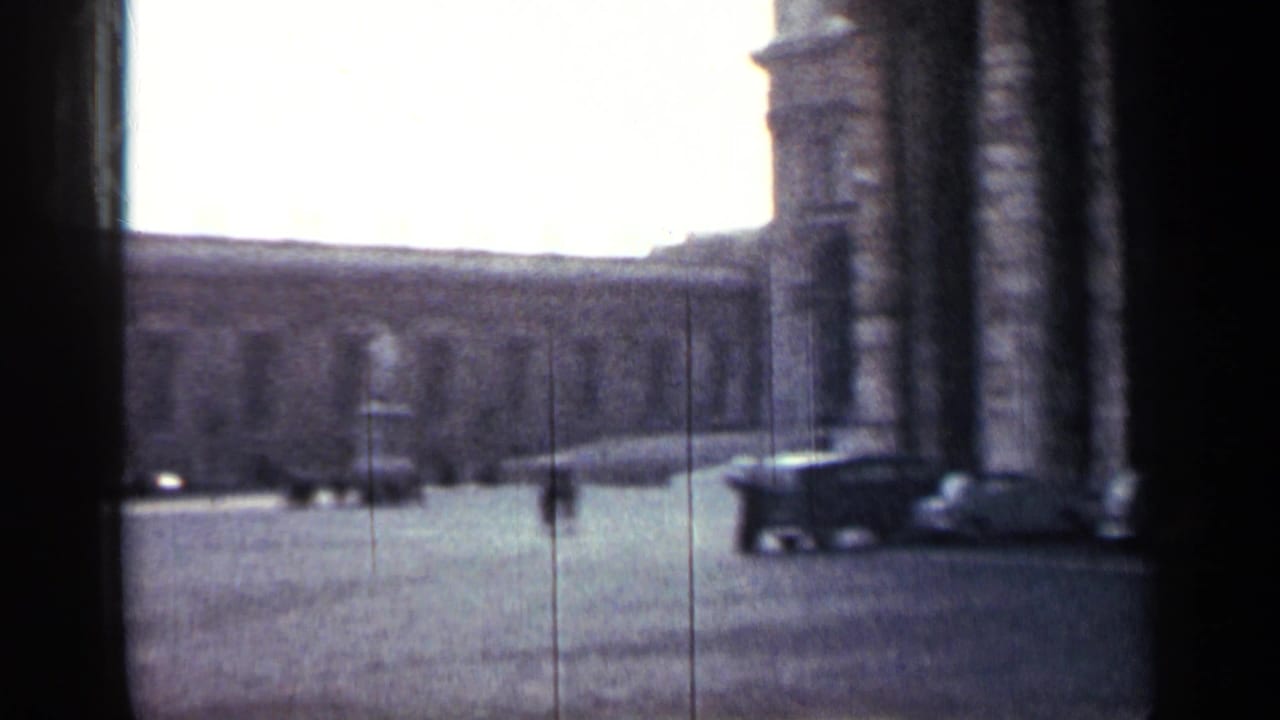 Columned building edge beside a broad paved plaza with a few distant figures and vehicles.