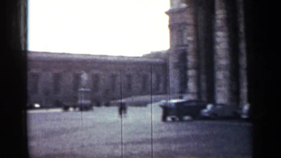 1962 Cuba 8mm frame showing a broad plaza beside large classical columns, with distant pedestrians and vehicles under bright daylight.