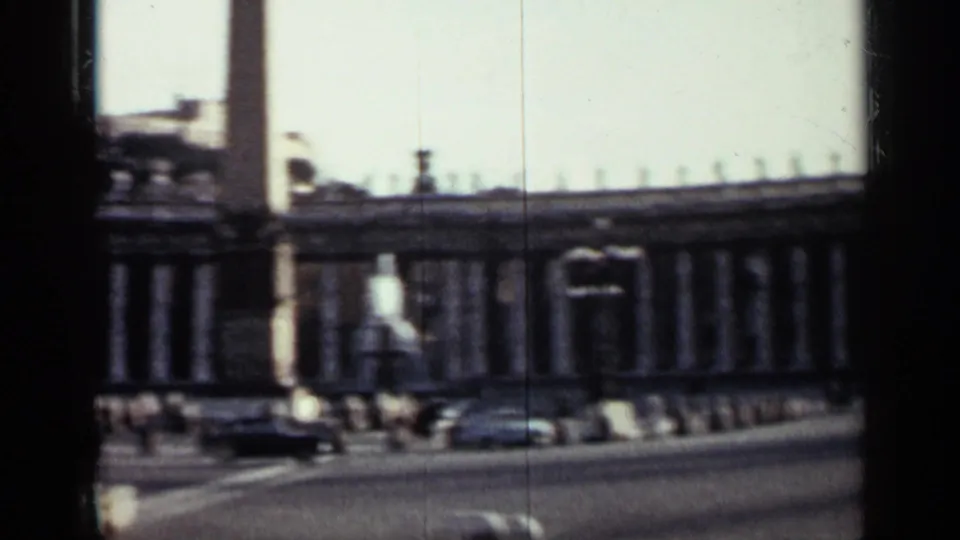1962 Cuba 8mm frame showing a columned plaza frontage, an obelisk-like monument, vintage cars, and distant pedestrians.