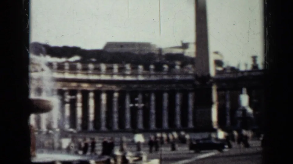 1962 Cuba 8mm frame with a wide view of the plaza, tall monument, classical colonnades, distant pedestrians, and street traffic.