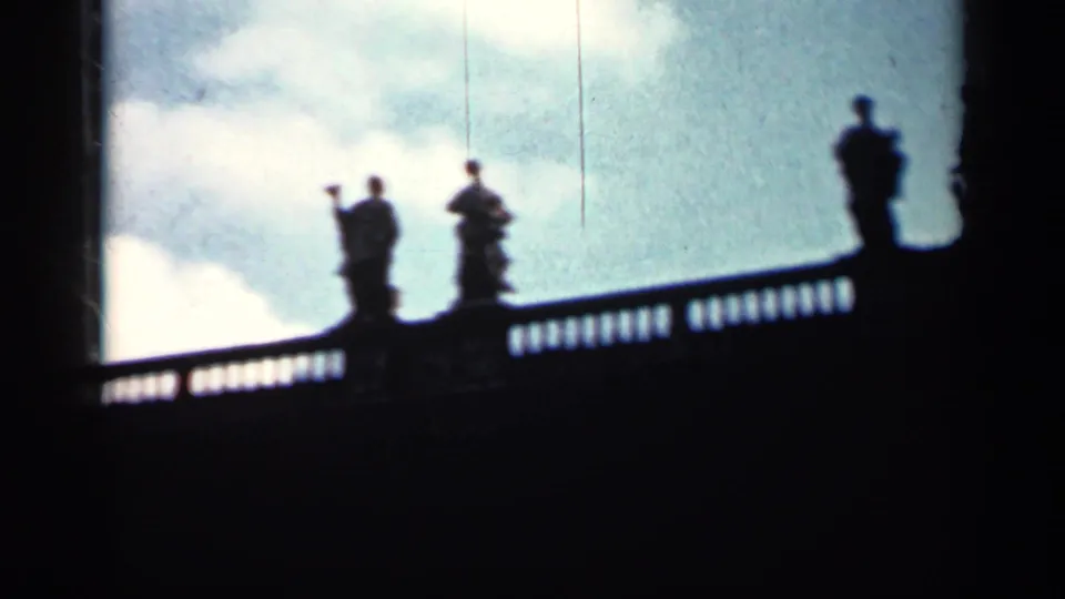 1962 Cuba 8mm frame showing dark statue silhouettes on a balustrade, with pale clouds and heavy shadow below.