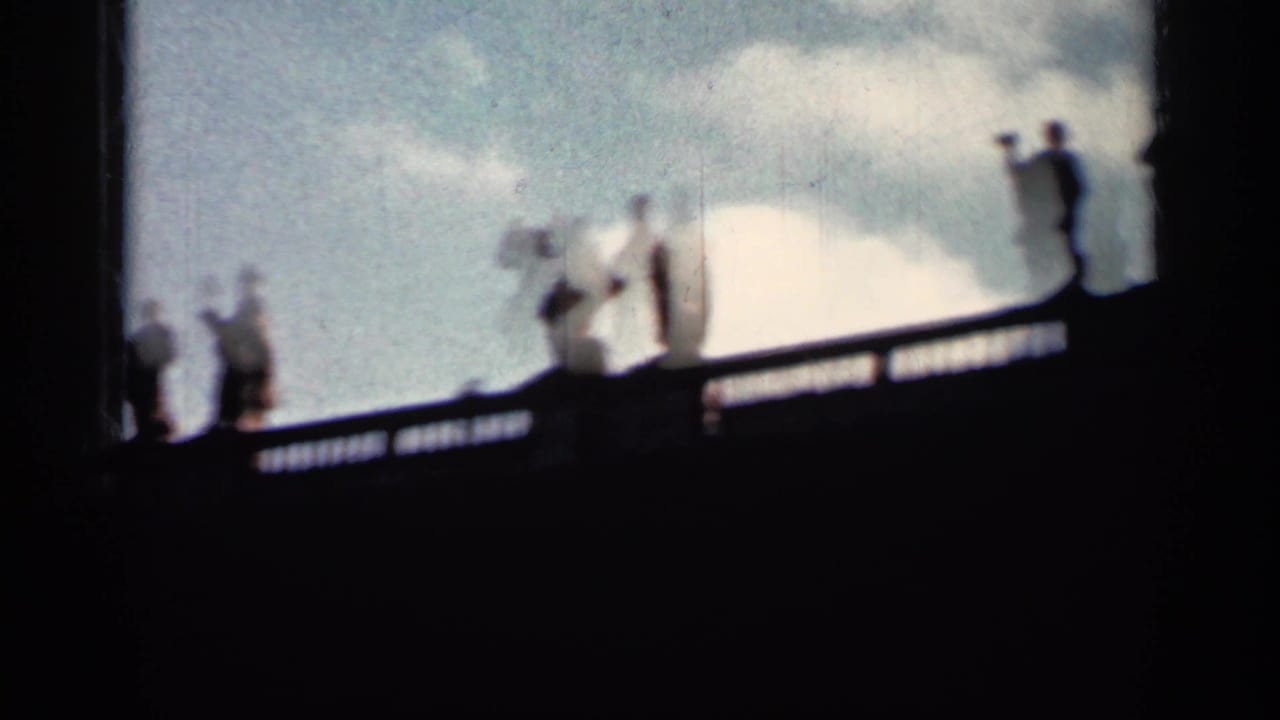 Silhouetted figures walk along a high balustrade against a bright cloudy sky.