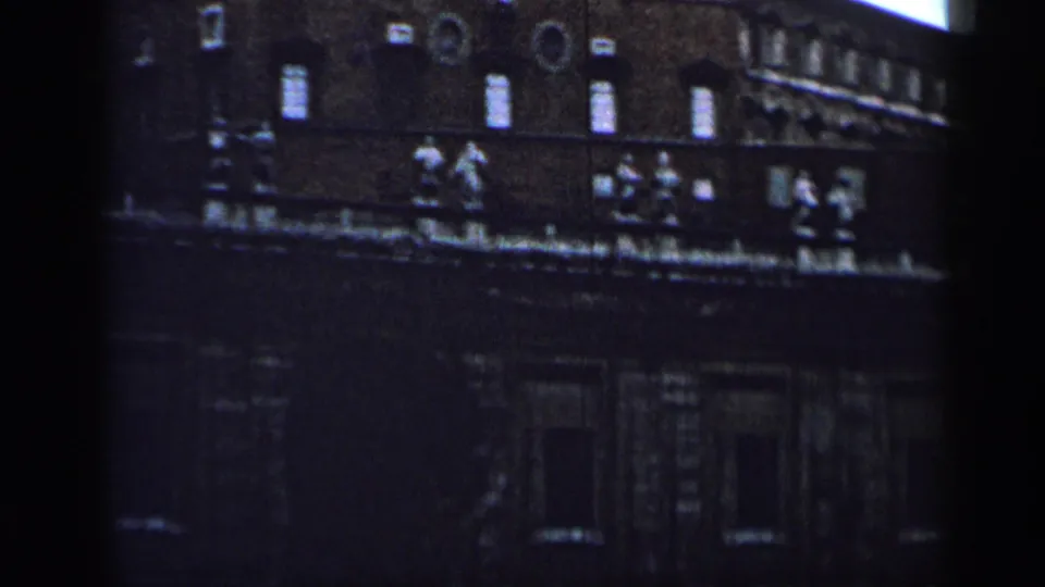 1962 Cuba home-movie frame showing a dim masonry building facade with small windows, roofline figures, and deep shadow.