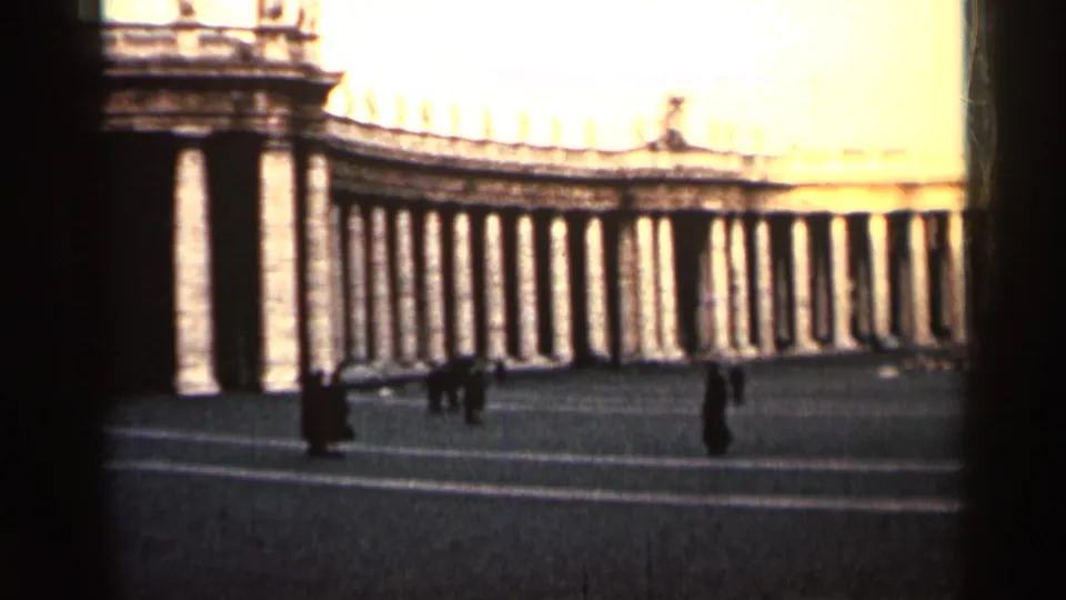 A sunlit colonnaded public plaza appears through soft 8mm focus, with small distant figures moving across the pavement.