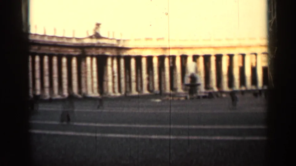 The 8mm frame shows a wide, sunlit colonnade and broad plaza, with very small people visible near the lower edge.