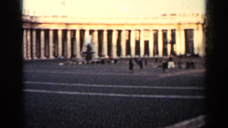 A warm, grainy 8mm frame shows a monumental colonnade, open paving, a fountain-like form, and distant visitors.
