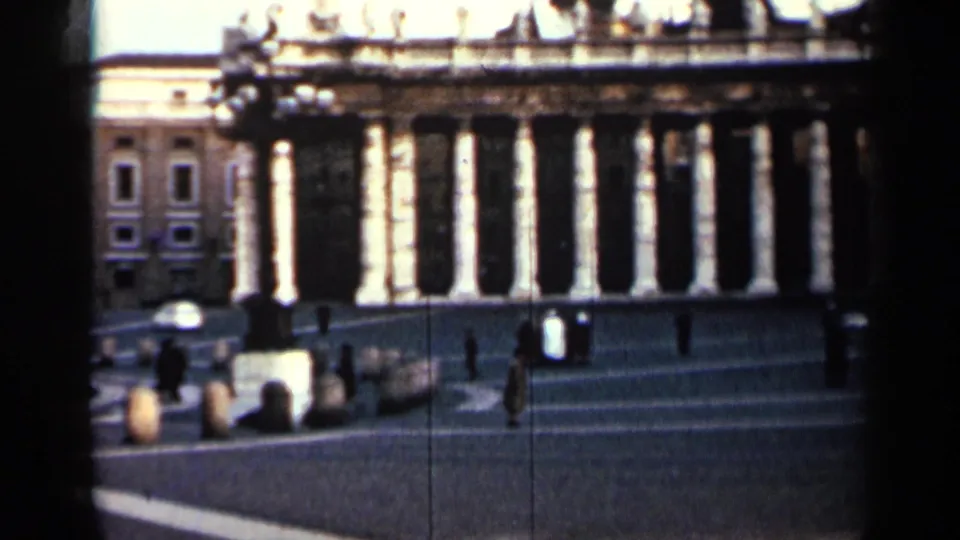 1962 Cuba 8mm frame of a monumental colonnade beside a plaza, with distant pedestrians and a fountain or monument in view.