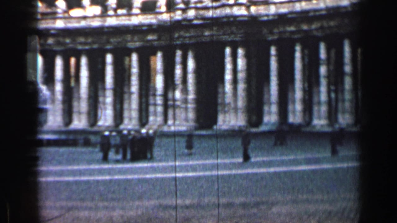 Colonnaded building facade across a wide plaza with small pedestrians in front.