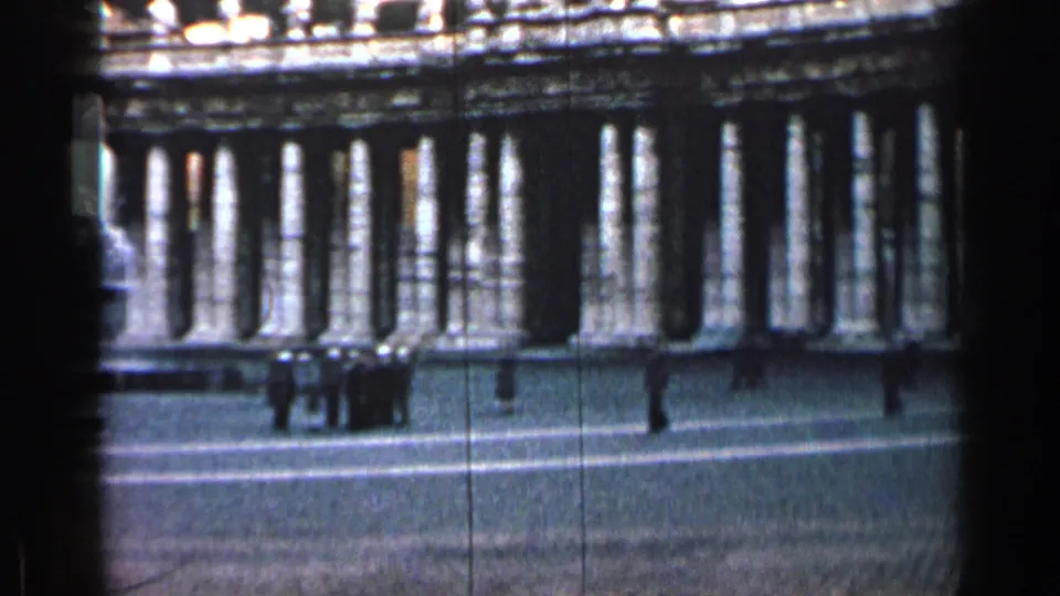 1962 Cuba home-movie frame showing a grand colonnade across a plaza with distant pedestrians and dark film-edge shadowing.