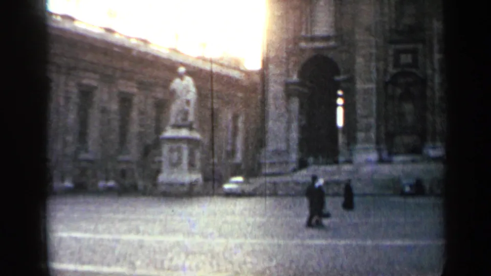 1962 home-movie view of an open plaza with a statue, pedestrians, a parked classic car, and a large arched doorway beside stone architecture.