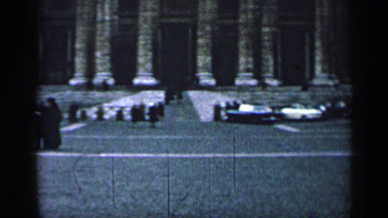 1962 Cuba home-movie frame showing a cobblestone street, parked cars, pedestrians, and tall columns on a grand stone facade.