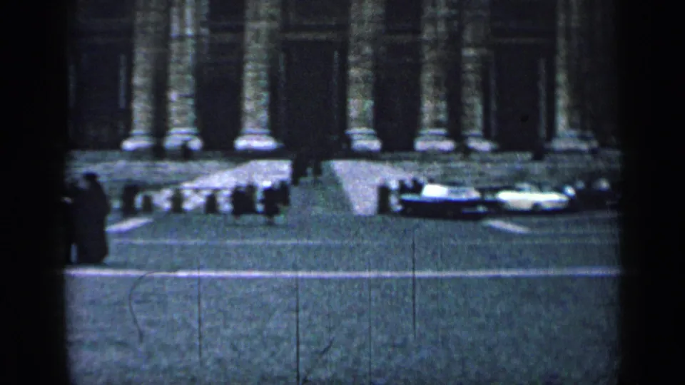 1962 Cuba home-movie frame showing a cobblestone street, parked cars, pedestrians, and tall columns on a grand stone facade.