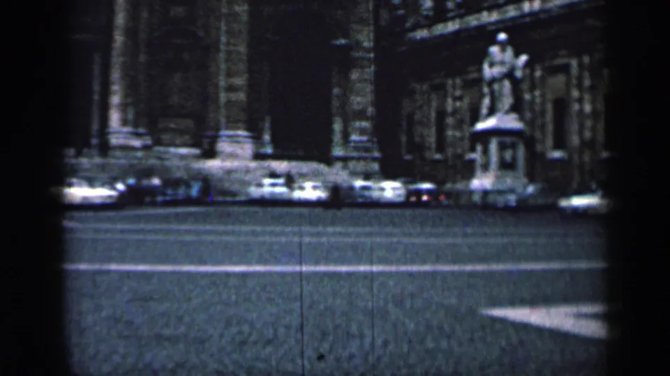 1962 Cuba street view with classic cars, a statue on a pedestal, and an ornate stone building seen through dark foreground obstruction.