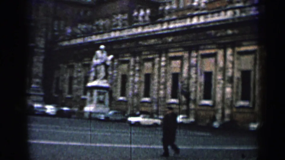 1962 Cuba frame showing a pedestrian crossing before a historic facade, statue, parked cars, and visible 8mm grain.