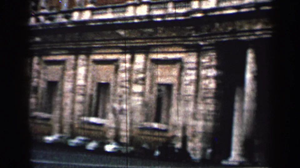 1962 Cuba home-movie view of a stone building facade with rectangular windows, parked cars, and a freestanding column near the edge.
