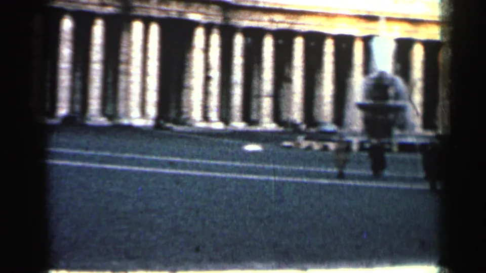 1962 Cuba frame with a distant pedestrian moving past a colonnaded facade and parked cars.