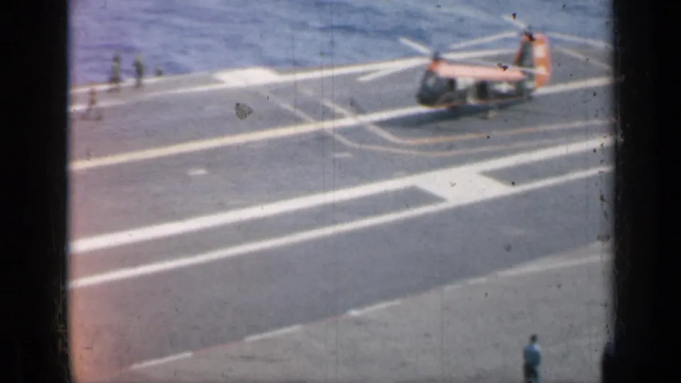 A small helicopter sits on an aircraft carrier deck with crew figures nearby and open water visible beyond the deck edge.
