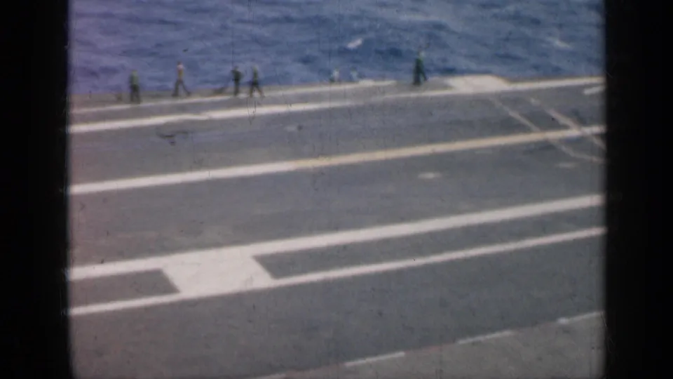 The carrier deck is seen from above with broad white markings, several distant crew members, and open sea behind them.