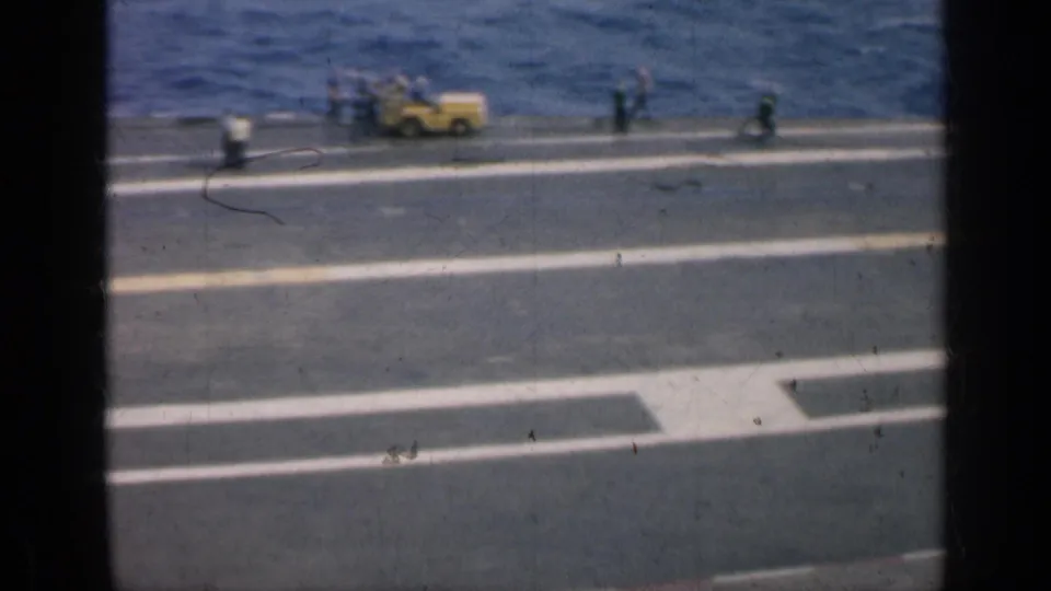 A yellow deck vehicle and several crew figures are visible on the aircraft carrier deck, with white markings and open water beyond.