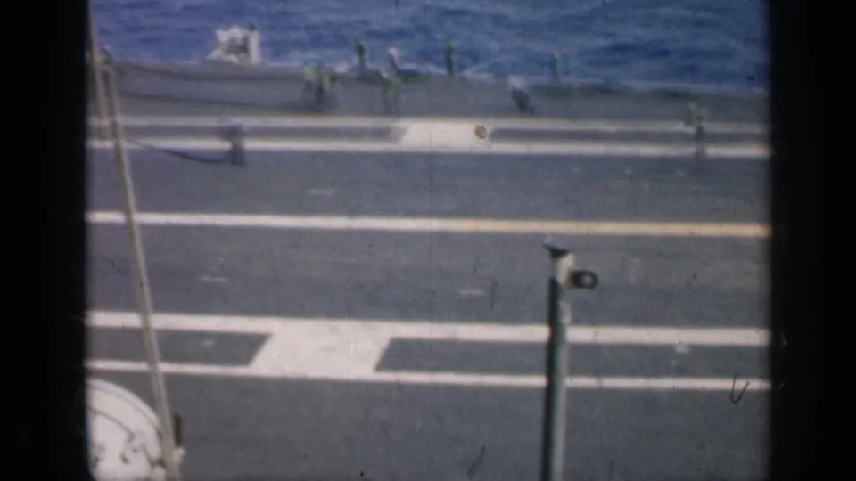 High-angle 1962 Cuba view of a carrier flight deck, with white markings, scattered crew, deck equipment, and ocean in the background.