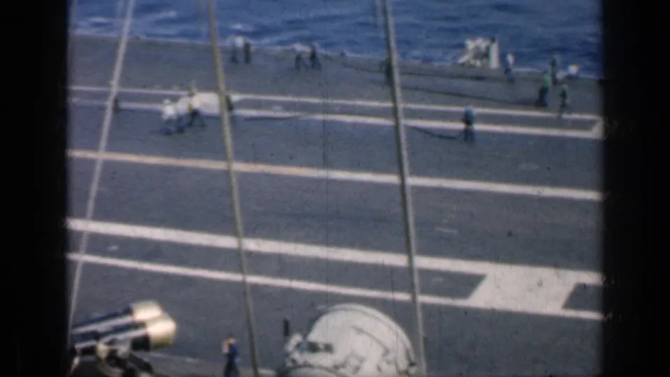 Elevated 8mm view from a carrier showing crew crossing marked deck areas, with rigging or cables in the foreground and ocean beyond.