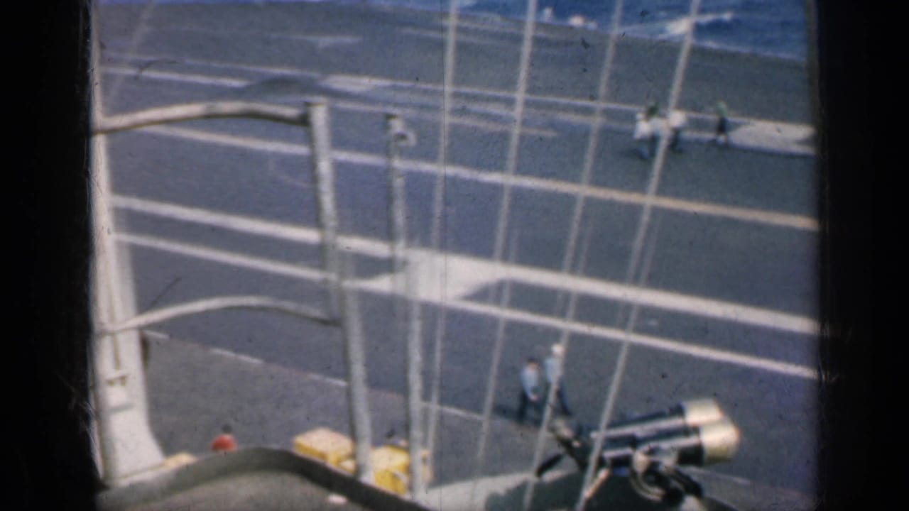 High view of an aircraft carrier deck with railings, deck markings, and small crew figures.