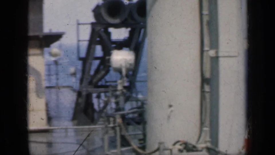 A mounted carrier deck device or aircraft-related structure appears beside railings and pale ship surfaces in grainy 8mm footage.