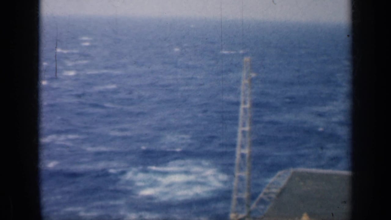 Carrier deck edge and mast over open ocean with a distant warship wake.