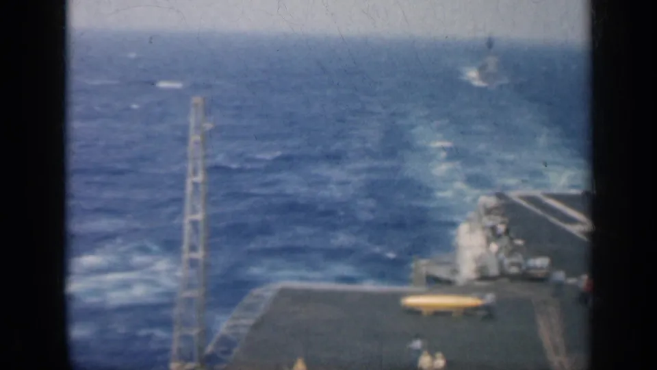 Aircraft carrier deck at sea near Cuba in 1962, with small crew figures, deck equipment and ocean wake behind the ship.
