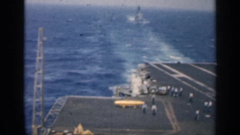 1962 Cuba footage showing a wider aircraft carrier deck view with crew scattered near equipment and a distant warship wake at sea.