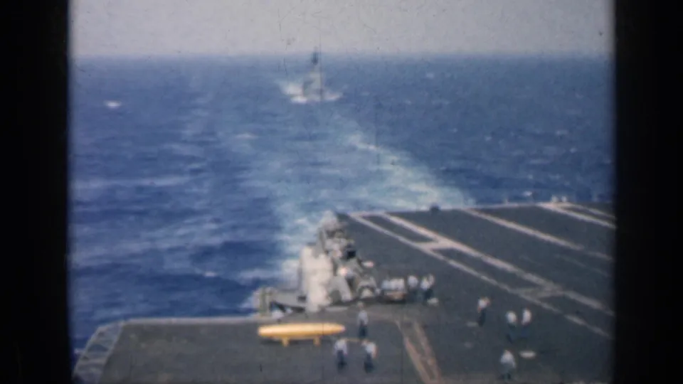 Aircraft carrier deck in 1962 Cuba footage, with crew near the deck edge and a distant warship wake visible across the sea.