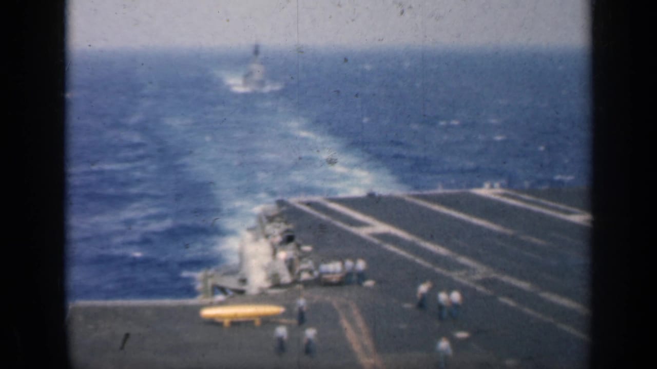 Aircraft carrier deck at sea with white wake, crew figures, and yellow equipment.
