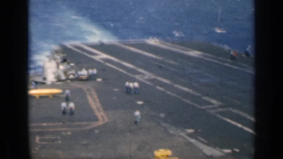 Large aircraft carrier flight deck underway at sea, with small groups of personnel visible against deck markings and surrounding blue water.