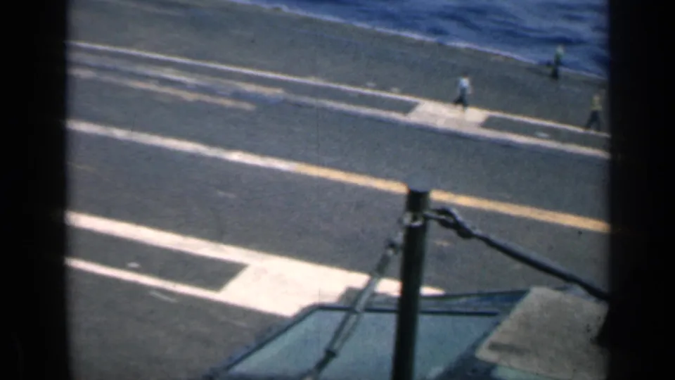 High-angle 1962 Cuba frame of a carrier deck at sea, with distant crew members, painted deck lines, and a foreground aircraft or helicopter surface.