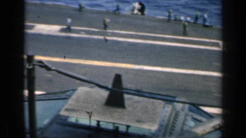 1962 Cuba carrier-deck frame with crew members moving across painted deck markings, open sea in back, and a partial aircraft or helicopter element below.