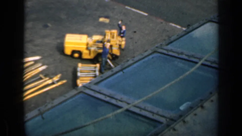 1962 Cuba home-movie frame showing a yellow deck utility vehicle, crew members, equipment, and partial aircraft or helicopter surfaces on a carrier deck.