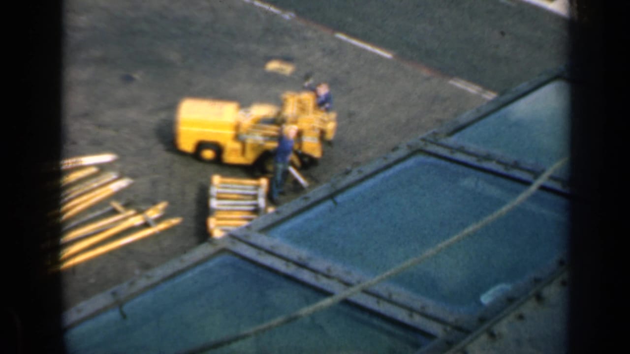 Elevated view of workers near a yellow industrial machine and stacked metal beams on pavement.