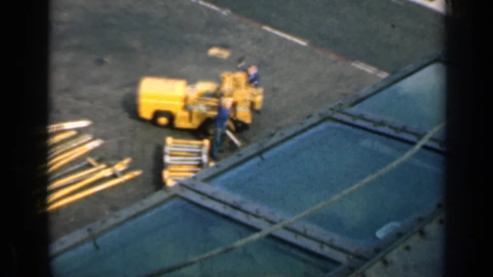 1962 Cuba 8mm frame showing workers around a yellow industrial machine with long metal beams on a paved site.