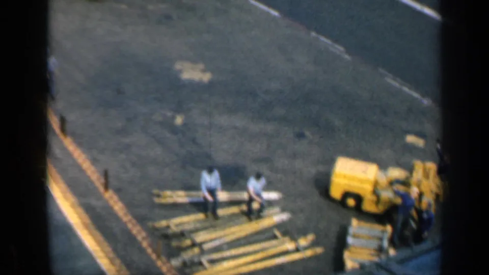 1962 Cuba 8mm frame with two workers by long beams and a yellow industrial machine at the edge of the shot.