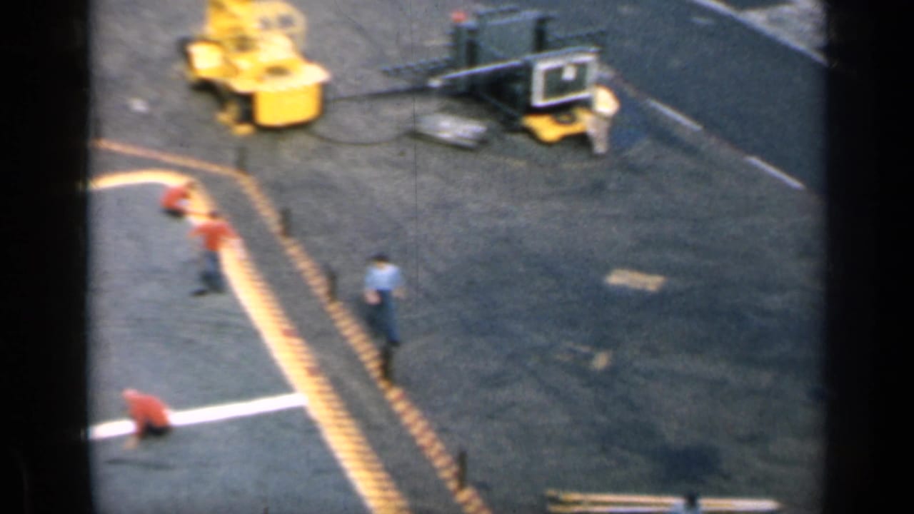 Aircraft carrier flight deck with yellow safety lines, deck crew, and wheeled equipment.