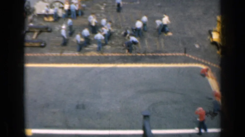 1962 Cuba-related 8mm frame with sailors and deck crew clustered near equipment on an aircraft carrier flight deck at sea.