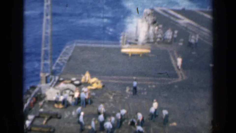 1962 Cuba-related 8mm frame showing sailors and carrier deck gear with ocean visible and a plume near the deck edge.