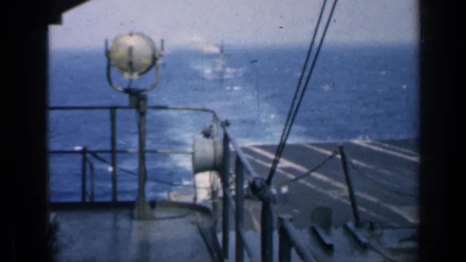1962 Cuba-related 8mm frame looking past a shipboard spotlight and rail toward the carrier deck edge, open sea, and distant vessel.