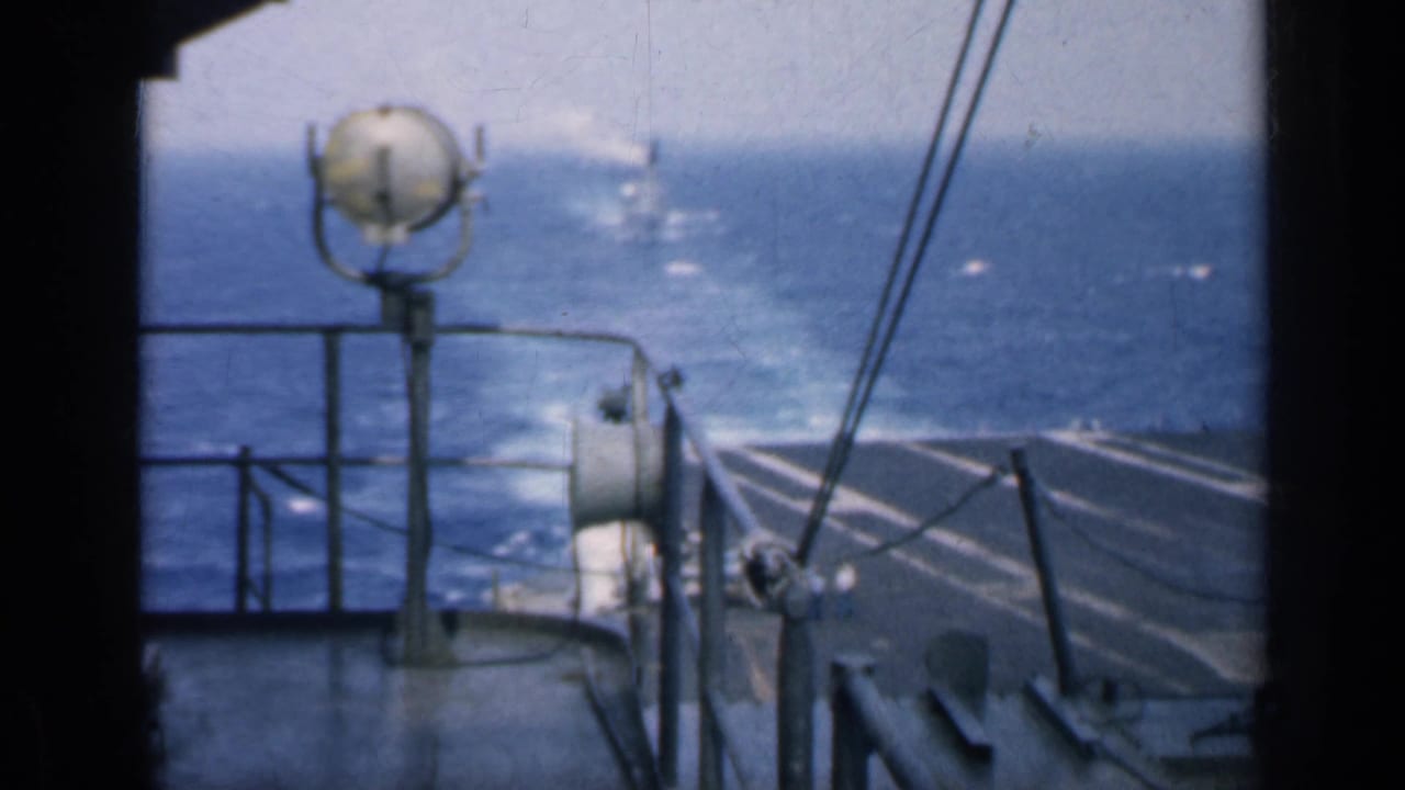 Carrier deck railings and a round shipboard fixture overlook open ocean with a distant vessel.