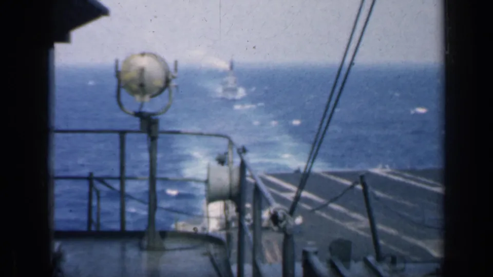 Carrier deck view over blue water, with railings, cables, a round shipboard fixture, and a distant vessel in hazy light.