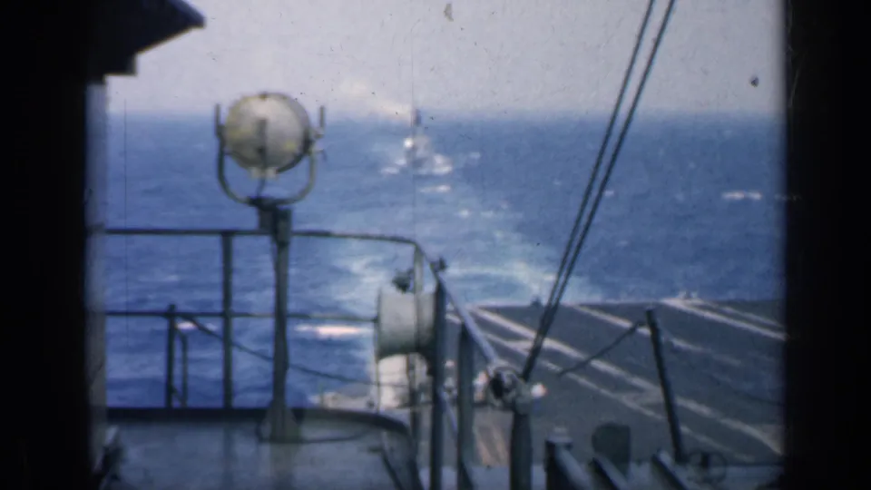 8mm carrier-deck perspective showing ocean, railings, deck equipment, and a distant vessel under a hazy horizon.