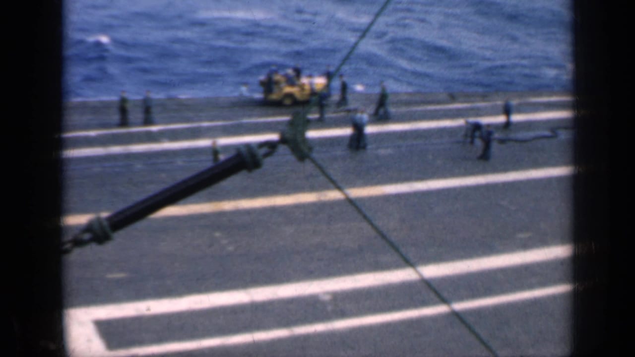 1962 Cuba-associated 8mm frame showing a U.S. aircraft-carrier deck, crew figures, ocean, and a yellow utility vehicle.