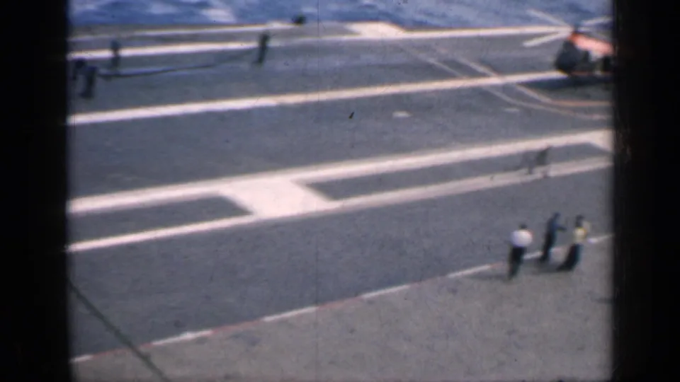 Carrier flight deck at sea with crew members, landing markings, and a helicopter visible in a 1962 Cuba-associated home-movie frame.
