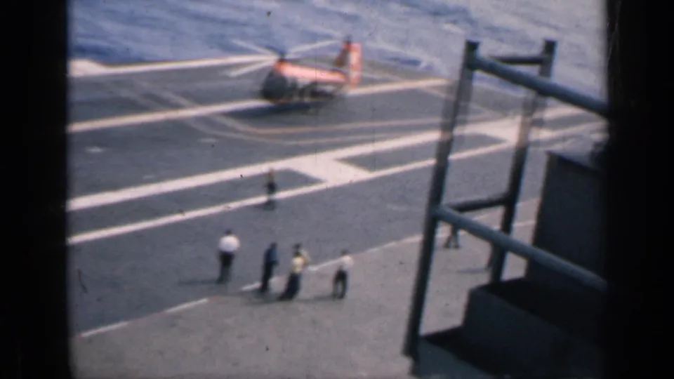 Handheld 8mm view of a U.S. aircraft-carrier deck near Cuba in 1962, with a helicopter, crew, railings, and sea visible.